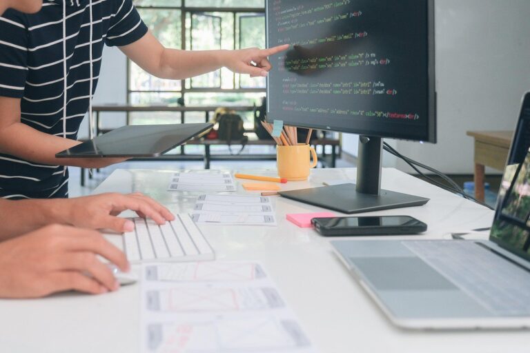 a photo of people at a desk with one person pointing to something on the screen of a computer and the other person typing